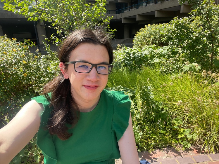 A selfie! I’m smiling at the camera, wearing a green dress, and sitting in front of a large amount of green foliage. It’s a sunny day and shining both on the side of my face and the plants.